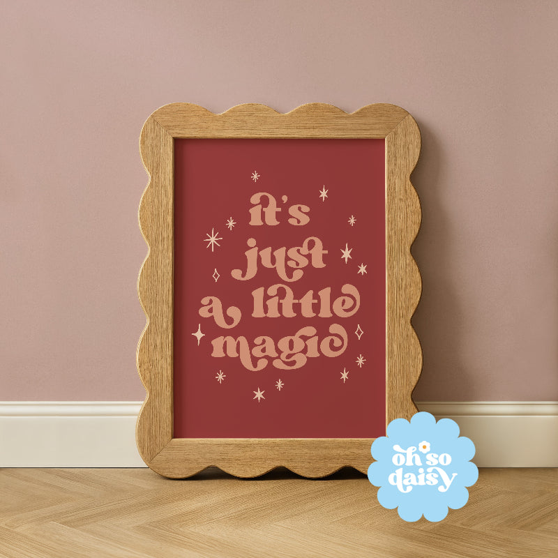 Wooden framed print with 'it's just a little magic' text on a red background, placed on a wooden floor against a pink wall.