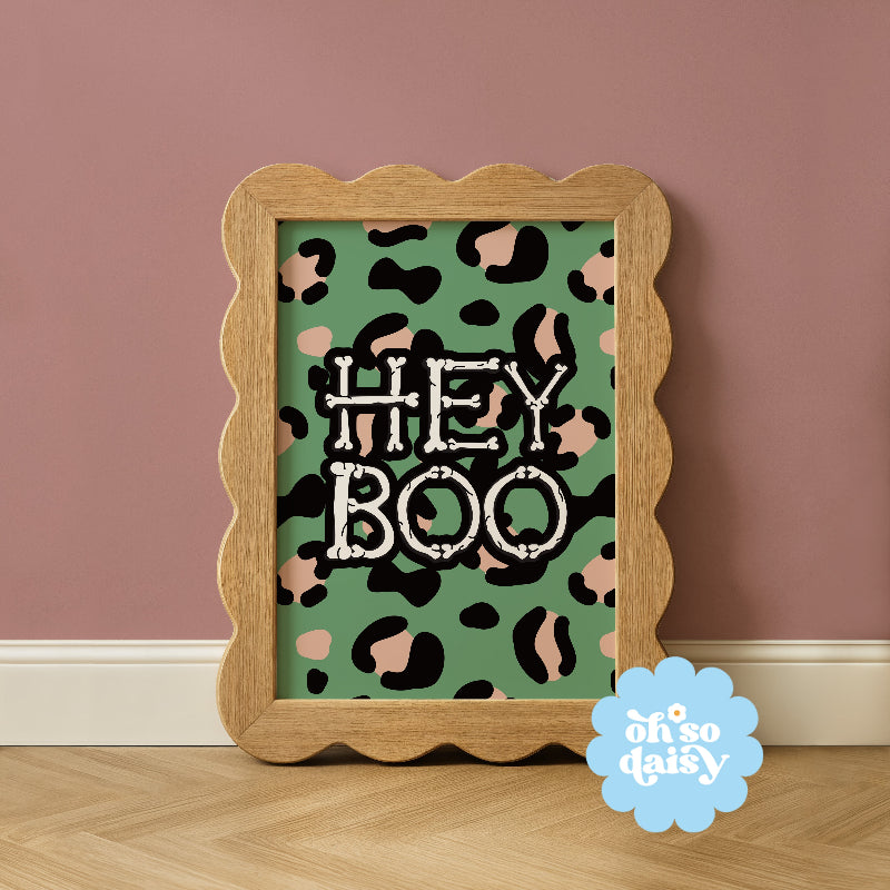 Framed artwork with 'HEY BOO' text on a green background with black and pink leopard print, against a pink wall.
