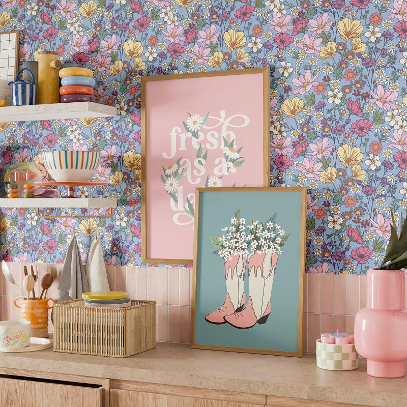 a kitchen counter with various decorative items, including a framed poster with a floral design, a pink and blue floral print, a pink vase, a basket, and a pink candle holder.