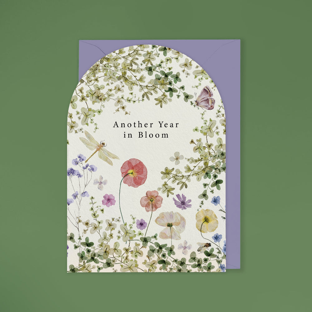 A floral greeting card with an arched top and the words 'Another Year in Bloom' centered on the front, shown with a lavender envelope behind it on a muted green background.