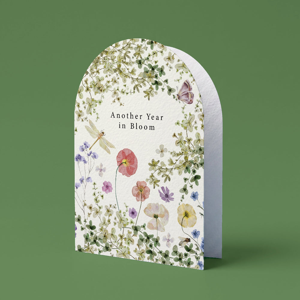 An arched greeting card with a white background and a delicate watercolor floral border, featuring the text 'Another Year in Bloom' and illustrated flowers, butterflies, and a dragonfly, set against a solid sage green background.