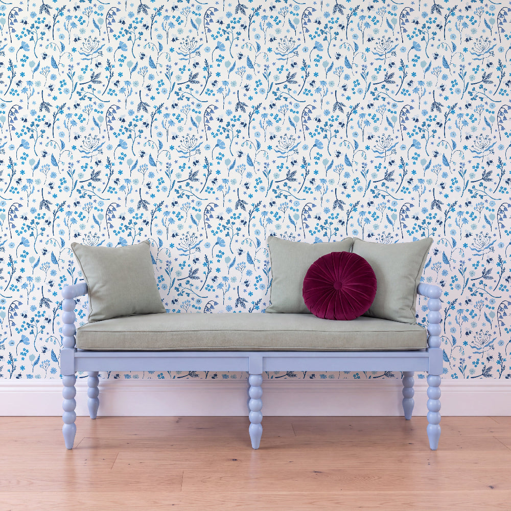 A light blue wooden bench with a pale sage green cushion and three pillows (two sage green, one deep magenta) set against a white wallpaper with a blue floral pattern, on a light wood floor.