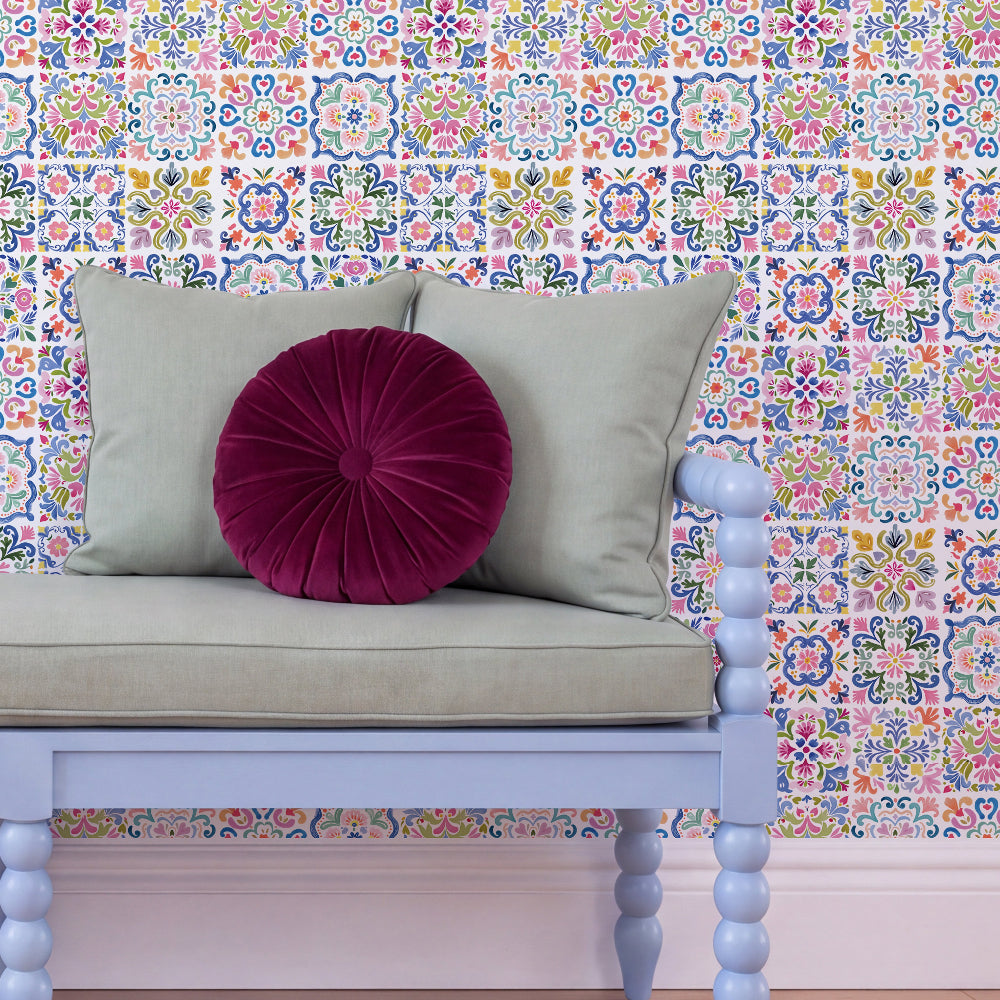 A light blue wooden bench with a beige cushion and two pillows, including a round deep magenta velvet accent pillow, set against a wall covered in colorful Moroccan-style patterned wallpaper.