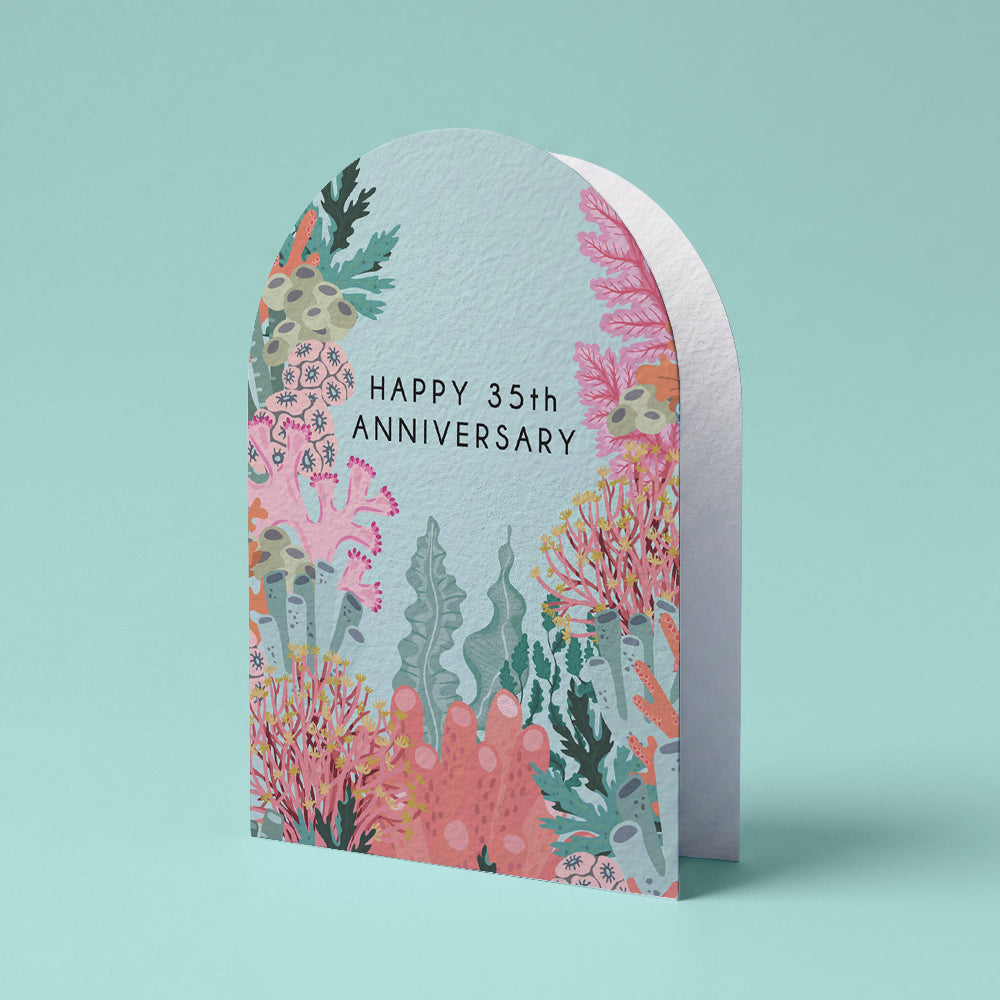 An arched anniversary greeting card with a coral reef illustration in pink, teal, and coral tones, featuring the text 'HAPPY 35th ANNIVERSARY' on a light aqua background, shown on a matching aqua backdrop.