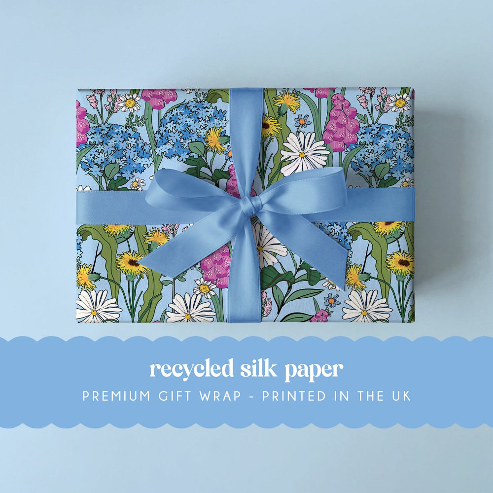 A rectangular gift box wrapped in light blue silk paper with a multicolor floral print, tied with a matching light blue ribbon and bow, on a pale blue background. A blue banner at the bottom reads 'recycled silk paper PREMIUM GIFT WRAP - PRINTED IN THE UK'.