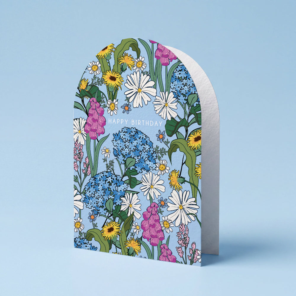 An arched birthday card with a colorful wildflower pattern on a light blue background, standing upright against a matching light blue backdrop. The card features white, blue, yellow, and pink flowers with 'HAPPY BIRTHDAY' printed in white near the top center.