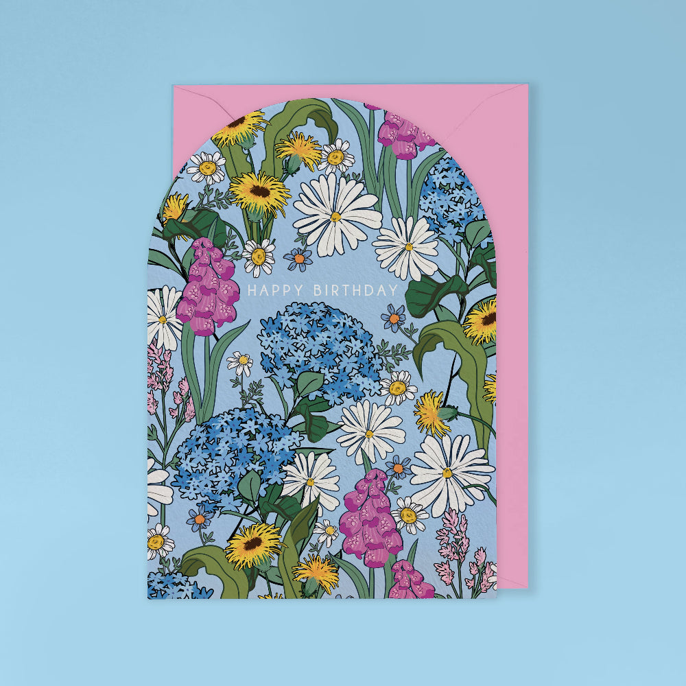 A floral arch-shaped greeting card with a pink envelope, featuring a dense pattern of white daisies, blue hydrangeas, yellow sunflowers, and pink blossoms on a light blue background, with 'HAPPY BIRTHDAY' printed across the center.