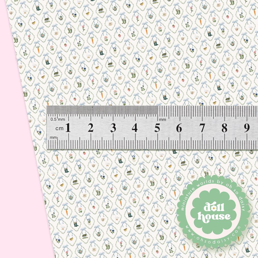 A close-up of a fabric or paper pattern swatch with a repeating small motif in blue, green, orange, and brown on a light background, with a metal ruler across the center for scale and a pink side panel. A green circular 'doll house' logo appears in the lower right.