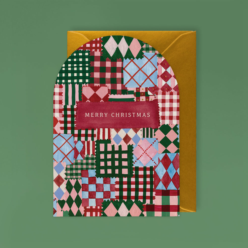 A Christmas greeting card with a colorful patchwork pattern in green, red, pink, and blue, featuring a gold envelope behind it on a solid green background.
