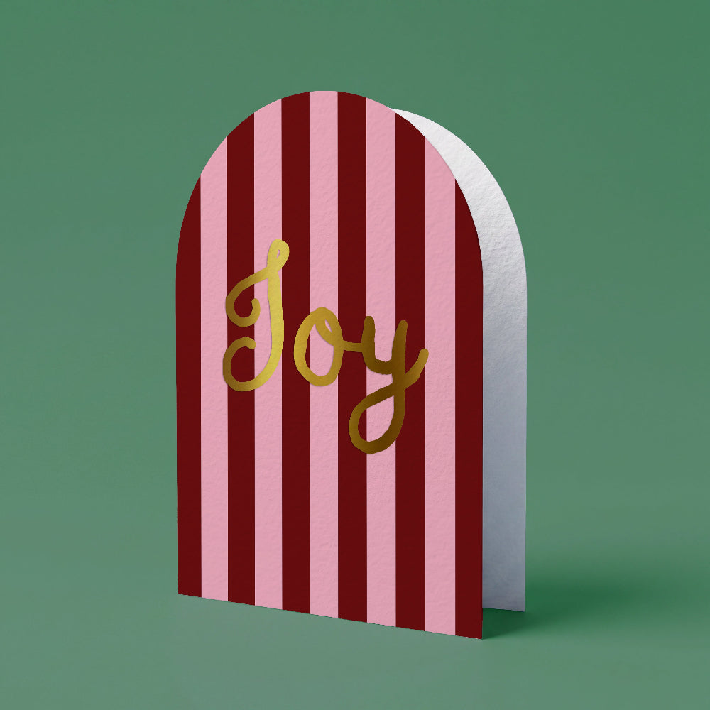 A festive arch-shaped greeting card with vertical pink and dark red stripes, featuring the word 'Joy' in metallic gold script, set against a solid green background.