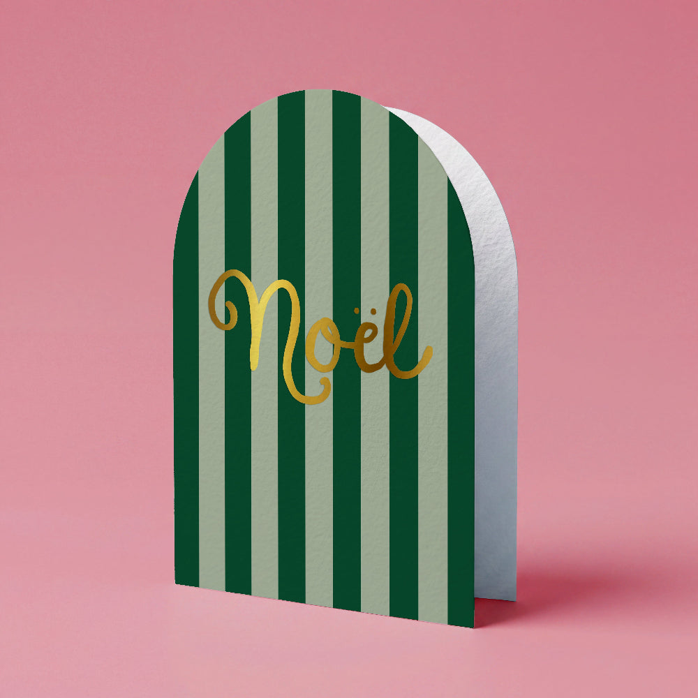 A festive arched greeting card with vertical dark green and light sage green stripes, featuring the word 'Noël' in metallic gold script, set against a solid pink background.