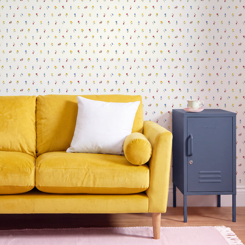 A yellow velvet sofa with a white throw pillow and a matching yellow cylindrical pillow, set against a wall with colorful vertical raindrop wallpaper. A dark blue metal side cabinet stands to the right on a light wood floor with a pink rug.