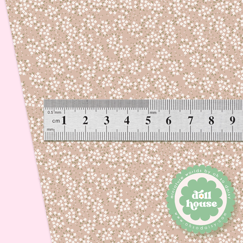 A close-up of a fabric sample with a beige background covered in a dense white floral print, shown with a metal ruler for scale and a green circular logo in the lower right corner.
