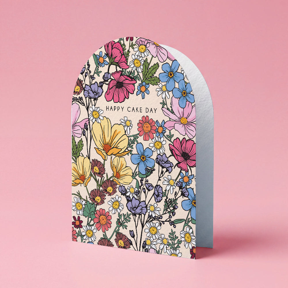 An arched greeting card with a colorful floral pattern and the words 'HAPPY CAKE DAY' printed on the front, standing upright against a solid pink background.