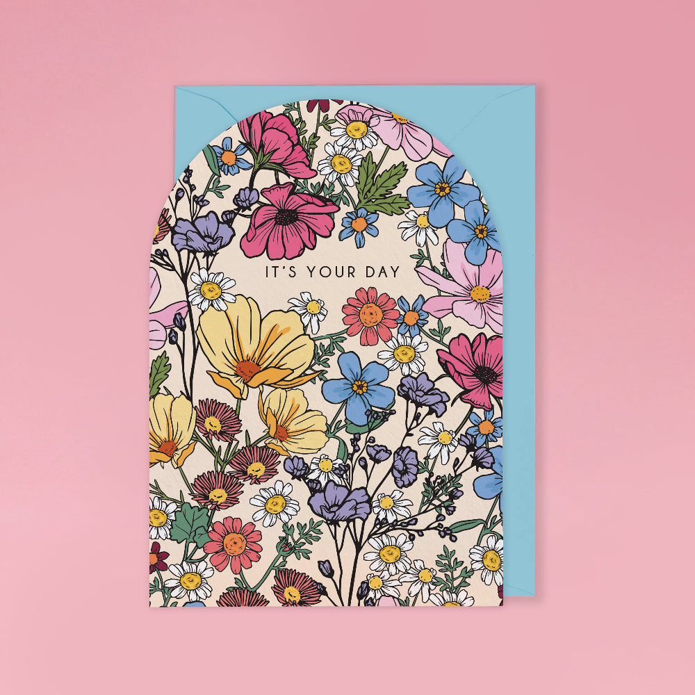 A floral greeting card with an arched top and the words 'IT'S YOUR DAY' in black text, layered over a light blue envelope on a solid pink background.