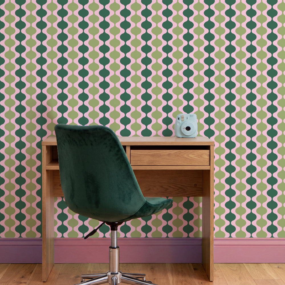 A styled home office corner featuring a mid-century modern wallpaper with a repeating green and light green geometric pattern on a dusty pink background. In front of the wall is a light oak wood desk with a single drawer, paired with a dark green velvet swivel chair and a small light blue bear figurine on top.
