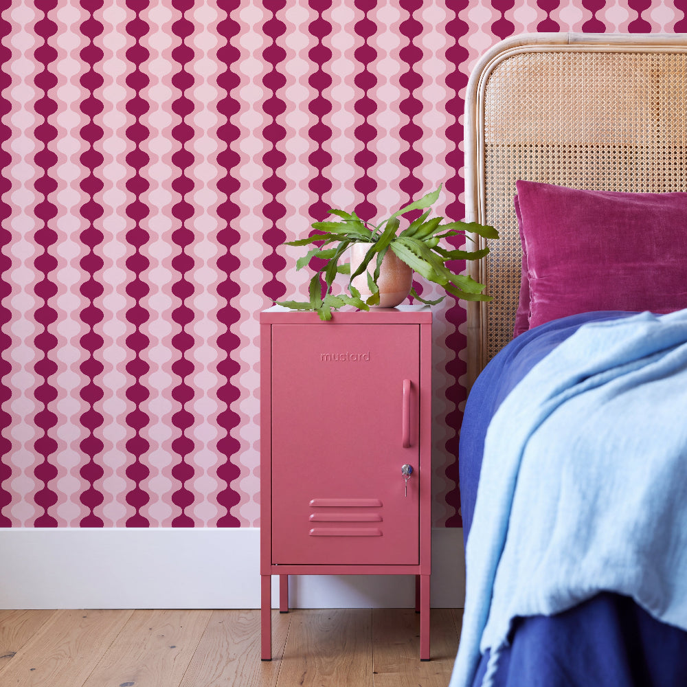 A pink metal nightstand in a styled bedroom setting, topped with a green potted plant. The background features bold magenta and pink vertical patterned wallpaper, a light wood cane bed headboard, and bedding in blue and magenta.