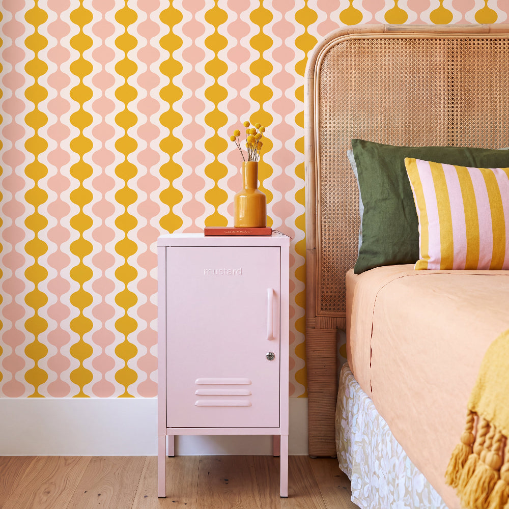 A styled bedroom corner featuring a light pink metal nightstand with a pink handle, set against a pink and yellow geometric patterned wallpaper. A small yellow vase with dried stems sits on top of the nightstand, which is positioned next to a bed with a woven cane headboard, green and striped pillows, and peach-colored bedding.