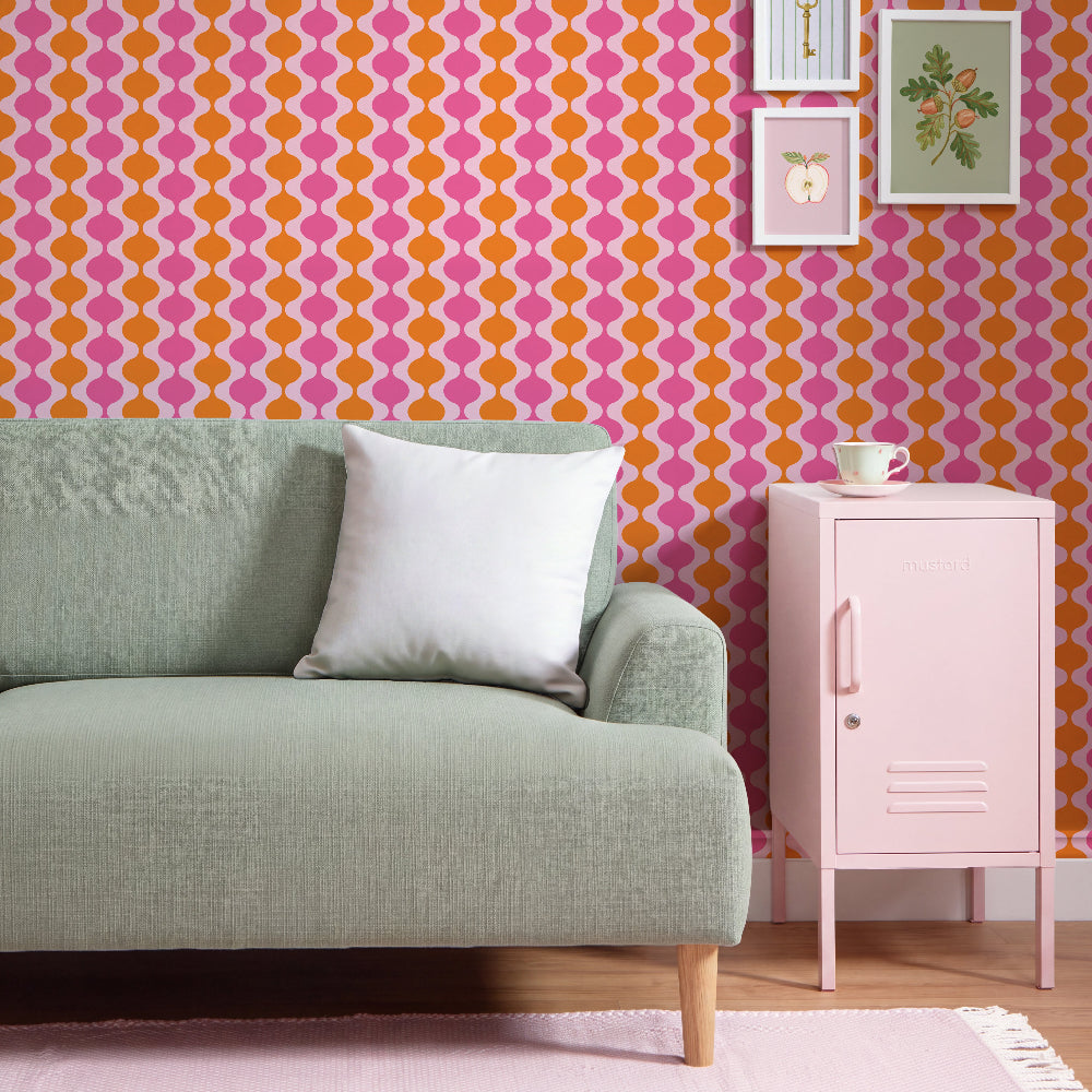 A styled interior scene with a mint green sofa, a white throw pillow, a pale pink side cabinet, and a bold pink-and-orange geometric wallpaper on the wall. No people or animals are present.