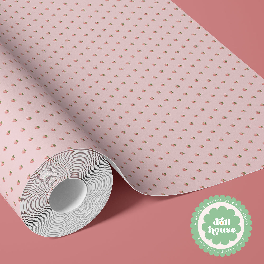 A roll of light pink wrapping paper with a repeating pattern of small pink and green strawberries, shown partially unrolled on a solid pink background. A green circular logo for 'đủ house' is visible in the bottom right corner.