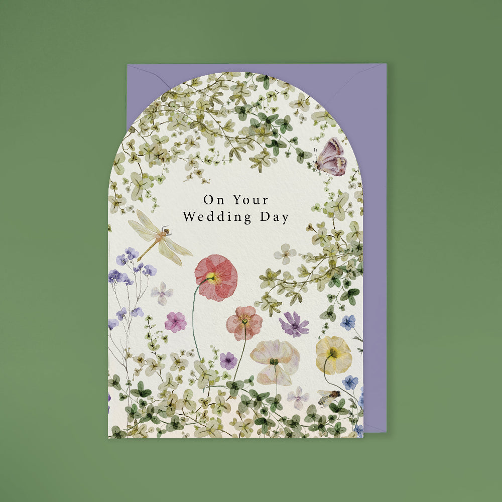 A floral wedding card with an arched top and the text 'On Your Wedding Day' in the center, shown with a lavender envelope against a muted green background.