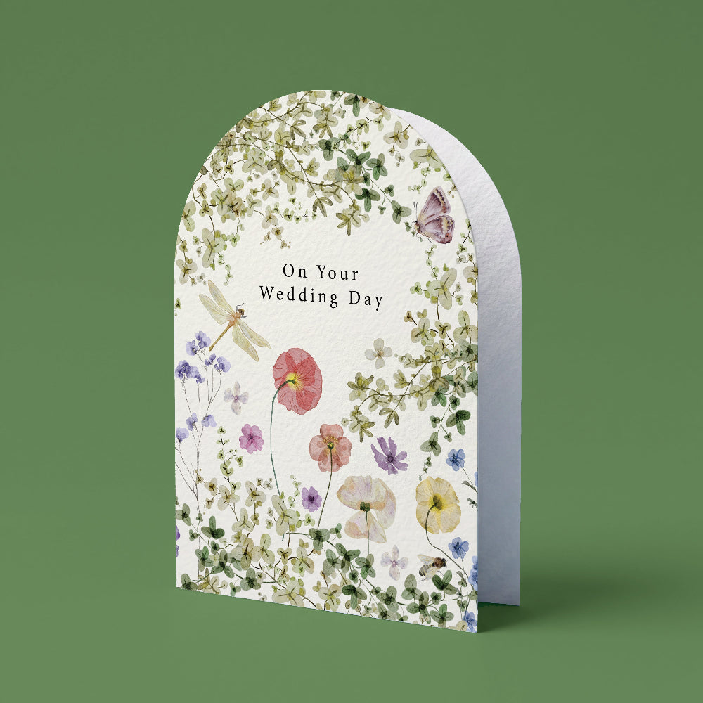 An arched greeting card with a white background and a delicate watercolor floral border, featuring the text 'Another Year in Bloom' and illustrated flowers, butterflies, and a dragonfly, set against a solid sage green background.
