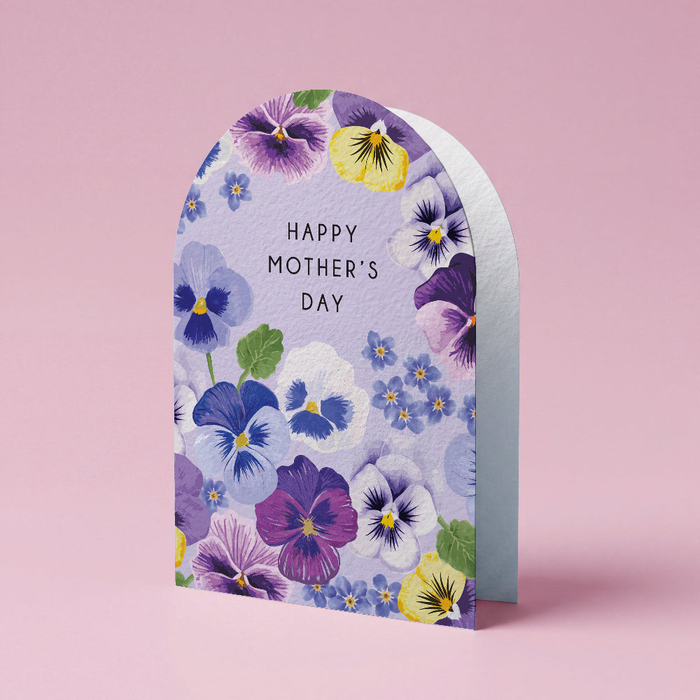 An arched Mother's Day greeting card featuring a watercolor floral design of purple, blue, and yellow pansies on a light lavender background, with 'HAPPY MOTHER'S DAY' printed in black. The card is shown standing upright on a solid pink background.