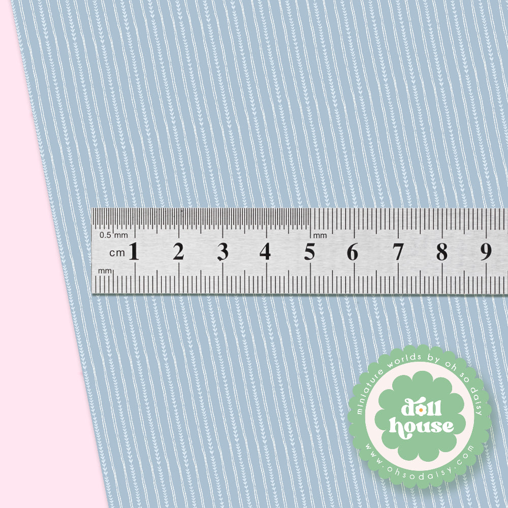 A close-up of a light blue and white striped fabric with a ruler showing the scale of the pattern, set against a pink background. A green 'doll house' brand logo is visible in the bottom right corner.
