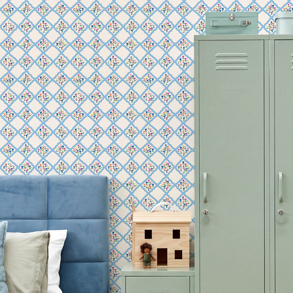 A styled bedroom corner featuring a blue tufted headboard, a small wooden house toy, a mint green locker, and a wall covered in blue and white diamond-patterned wallpaper with a small floral motif.