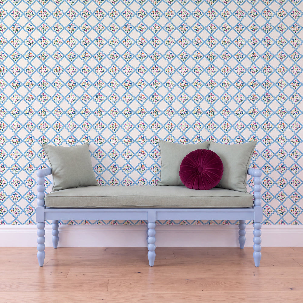 A light blue wooden bench with a pale sage green cushion and two pillows, including one deep magenta round pillow, set against a white wallpaper with a blue geometric pattern and light wood flooring.