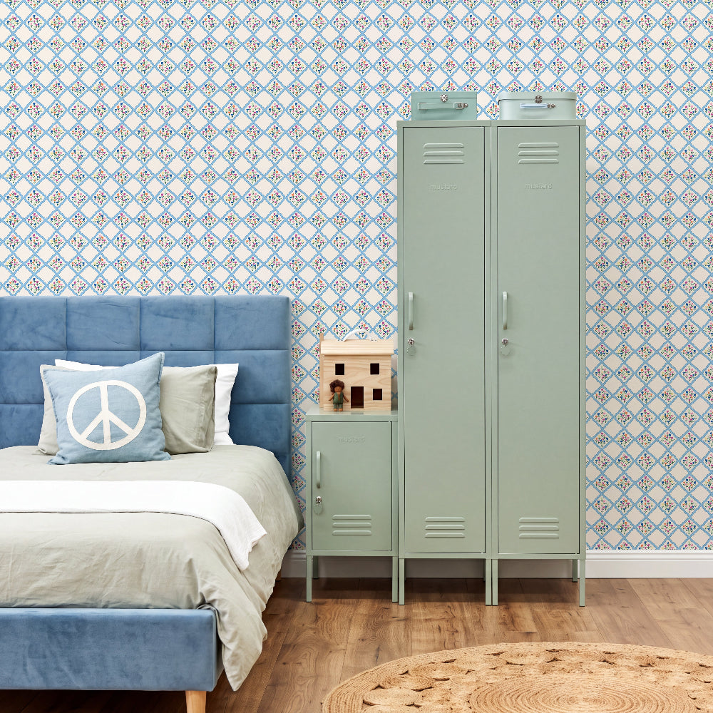 A styled bedroom scene featuring a tall mint green metal locker and matching nightstand next to a blue upholstered bed, set against a blue and white patterned wallpaper and a wood floor.