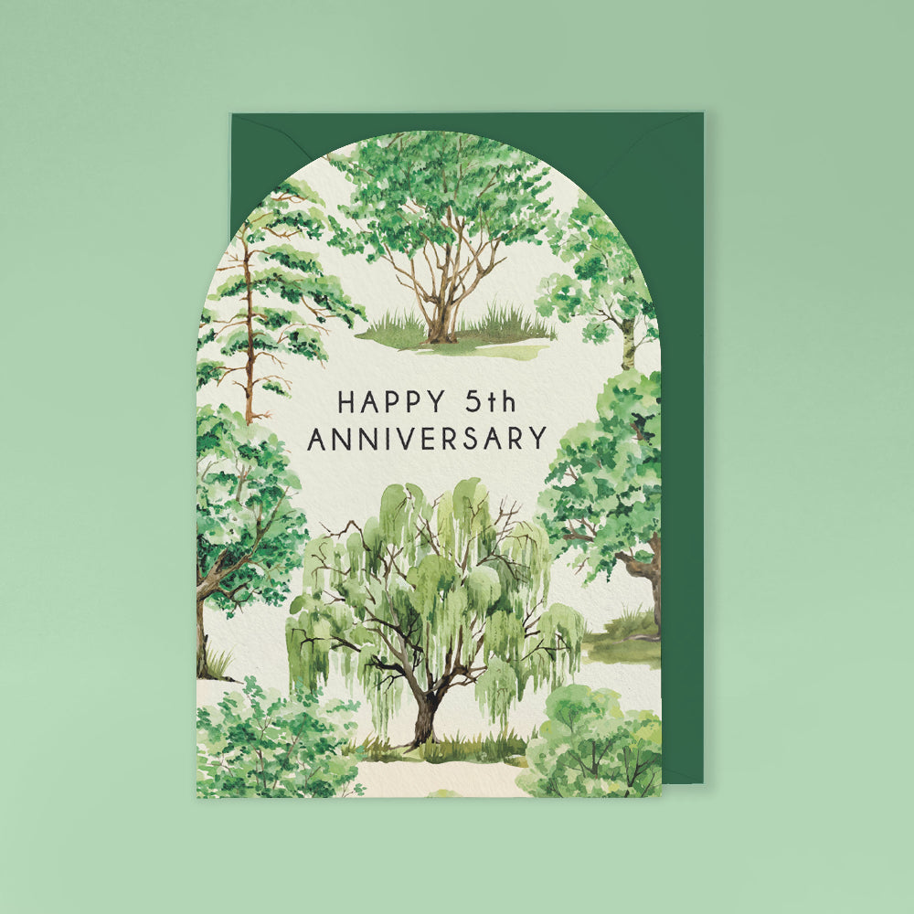 An arched anniversary card with a dark green envelope behind it, featuring a watercolor-style forest scene and the text 'HAPPY 5th ANNIVERSARY' in the center.