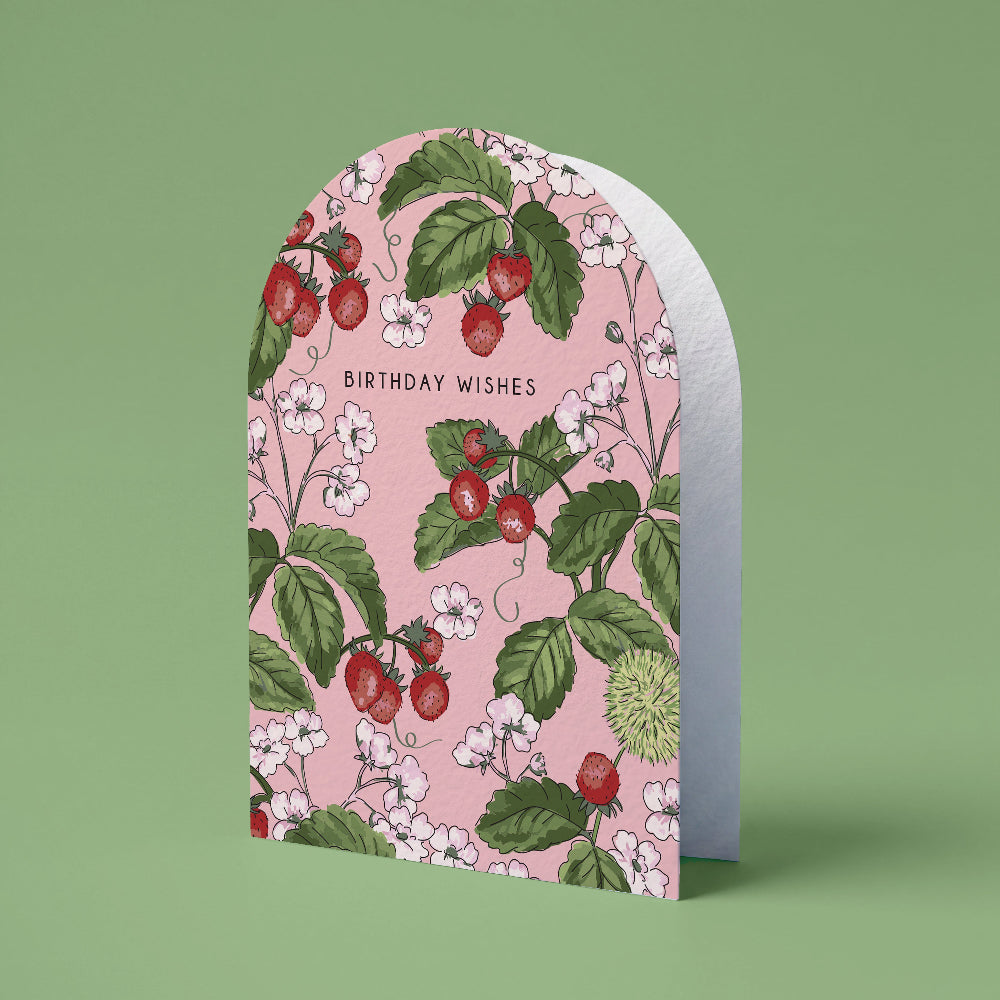A tombstone-shaped greeting card with a pink background, illustrated with red strawberries, green leaves, and white flowers. The card is displayed standing upright against a solid sage green background, with the text 'BIRTHDAY WISHES' printed on the front.