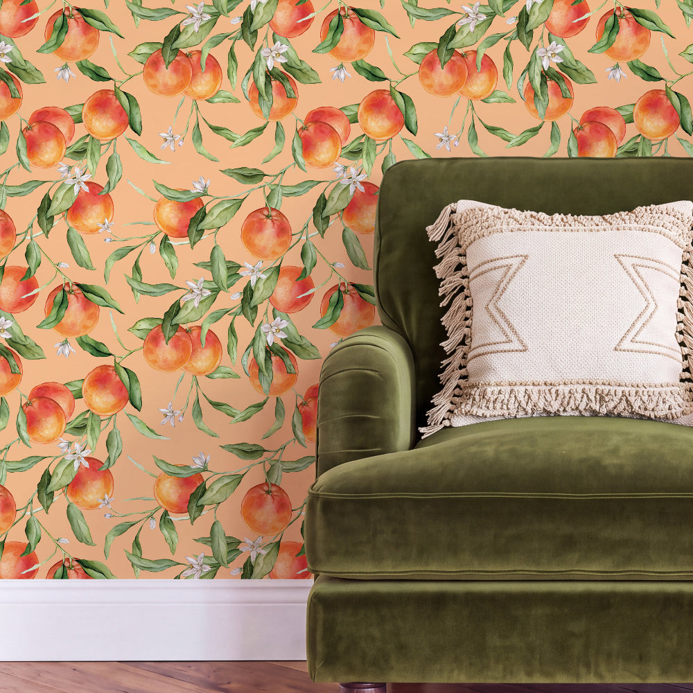 A styled interior scene showing a peach and orange botanical wallpaper with green leaves and small white flowers behind an olive green velvet armchair with a cream fringed throw pillow.
