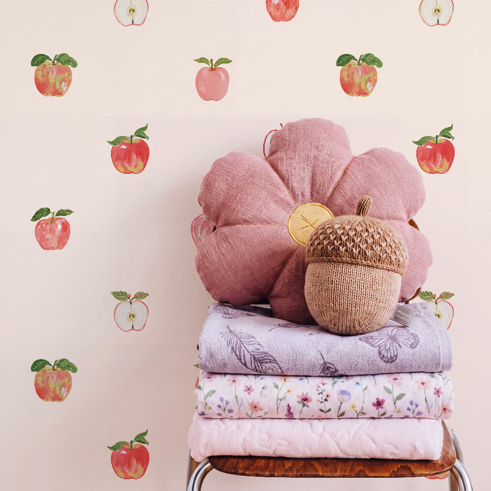 A styled interior scene showing a stack of folded pastel textiles on a wooden chair, topped with a large dusty pink flower-shaped pillow and a small acorn-shaped cushion. The wall behind is cream with scattered apple illustrations.