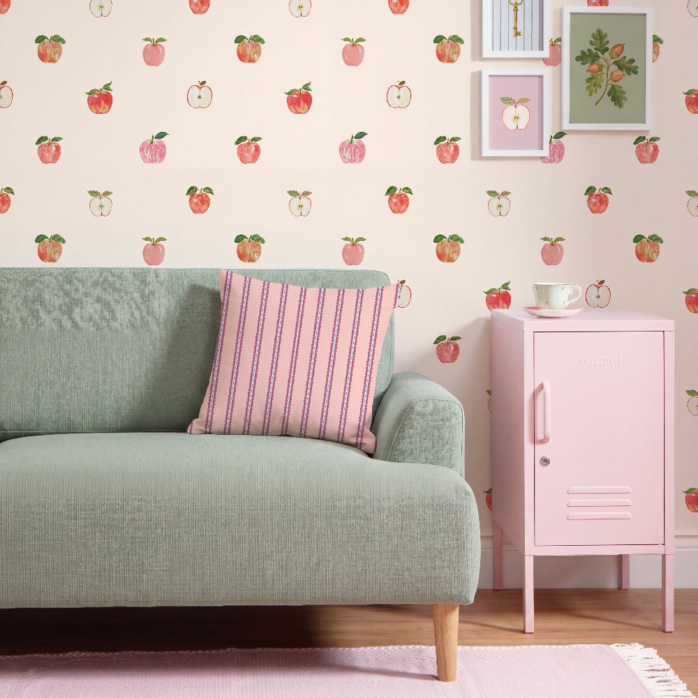 A styled interior scene with a pale green sofa, a pink striped throw pillow, a pink cabinet, and wallpaper covered in a repeating watercolor apple print on a light blush background.