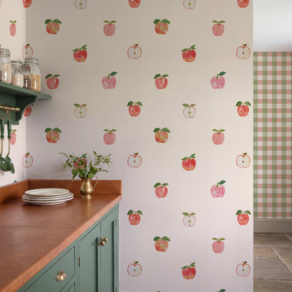 A kitchen interior featuring wallpaper with a repeating pattern of red and pink apples on a white background, paired with teal cabinets, a wood countertop, and a gingham-patterned wall panel in the adjacent room.