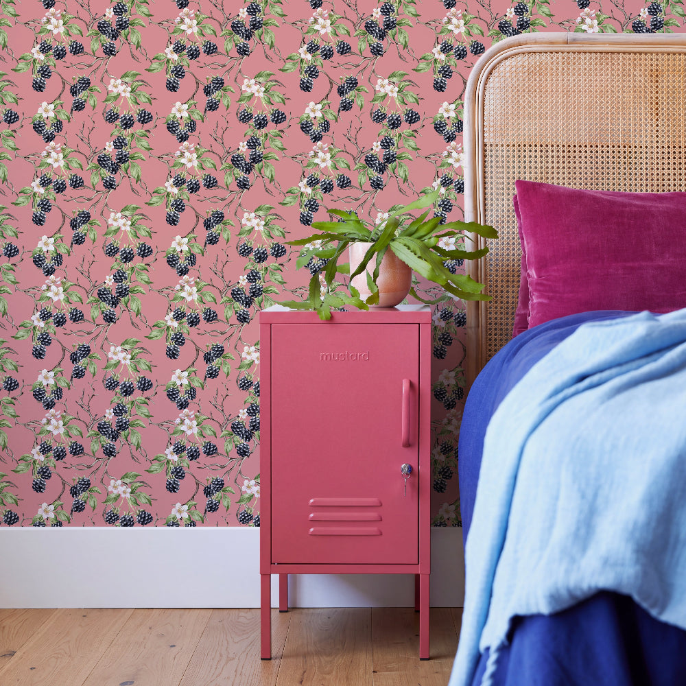 A styled bedroom corner featuring a pink metal nightstand with a plant on top, set against a pink wallpaper with a blackberry and white flower pattern. To the right is a bed with a blue blanket and a magenta pillow, and a woven cane headboard is visible.