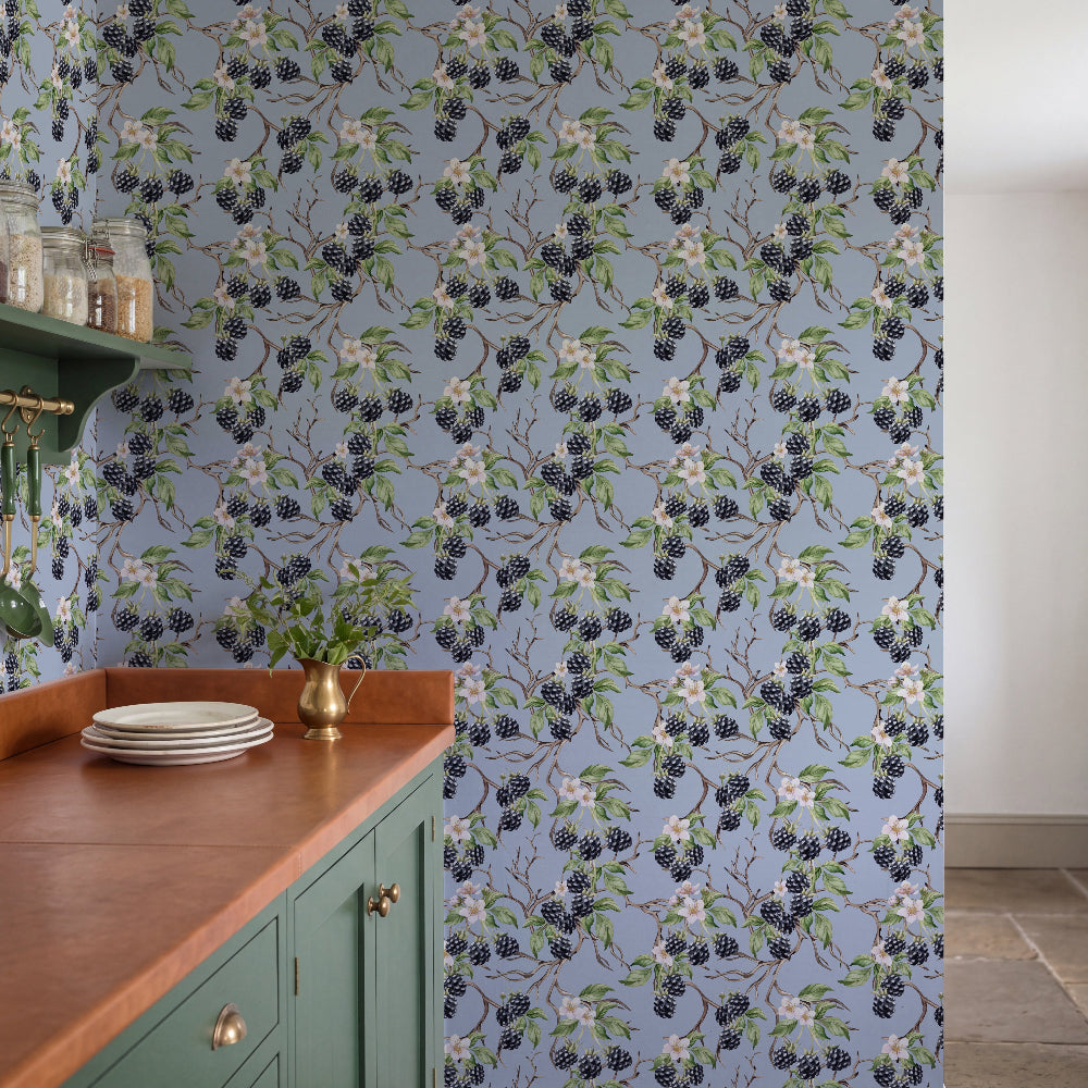 A kitchen interior featuring a light blue wallpaper with a repeating pattern of blackberries on branches, green cabinets, a wooden countertop, and a brass vase.