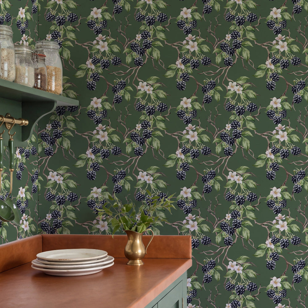 A kitchen scene featuring dark green floral and berry patterned wallpaper, a teal-green cabinet with a copper-wood countertop, white plates, and a small brass pitcher.