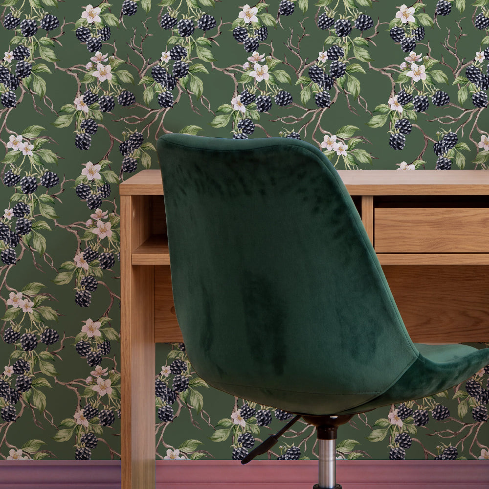 A dark green velvet office chair positioned in front of a light wood desk, set against a wall covered in dark green floral and berry patterned wallpaper.