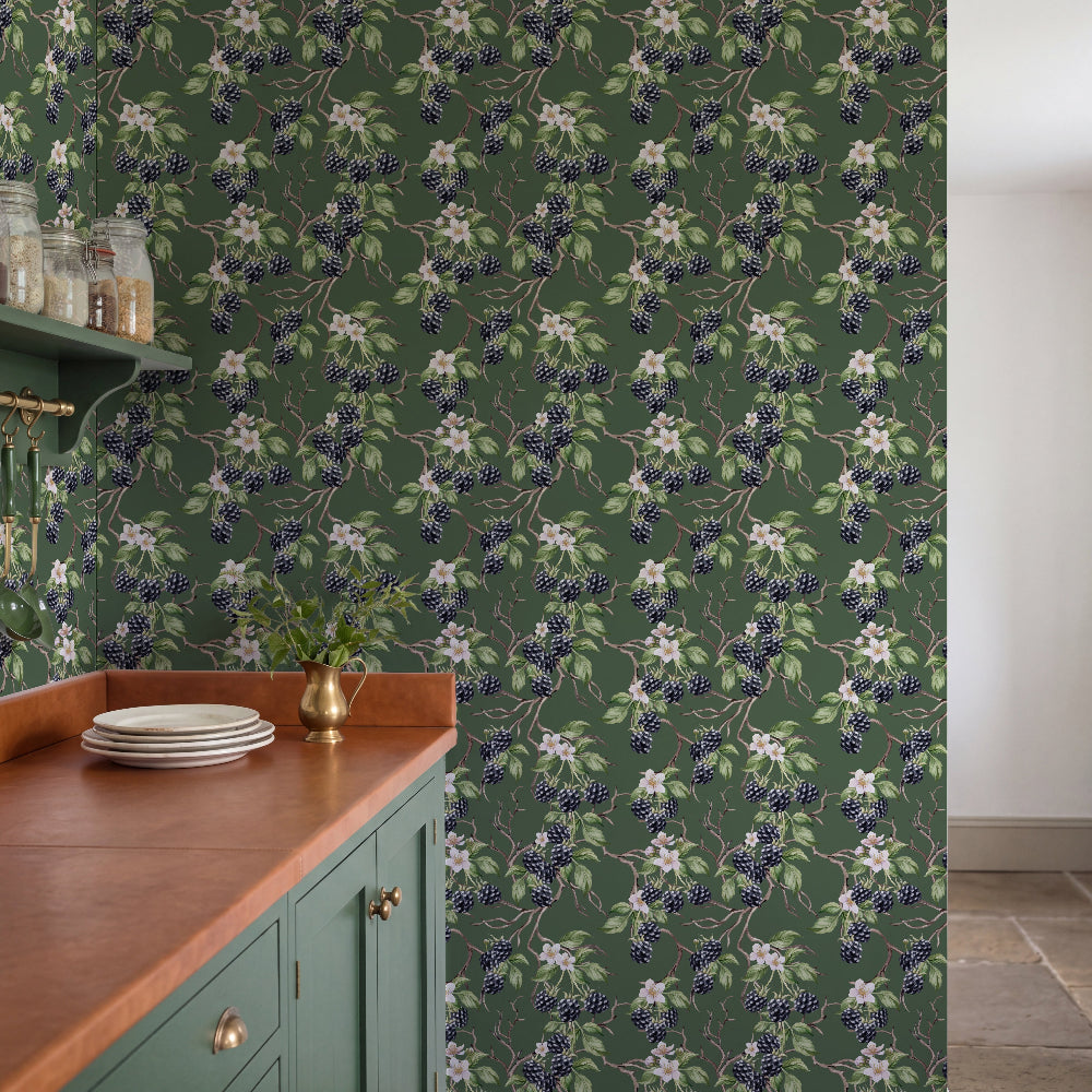 A kitchen interior featuring dark green wallpaper with a repeating floral and berry pattern, a wooden countertop, teal cabinets, and a brass vase with greenery.