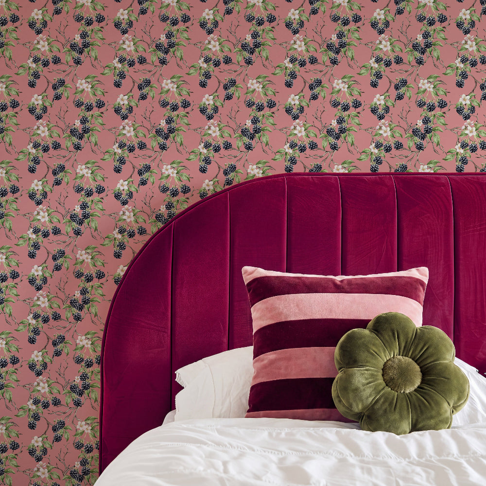 A styled bedroom corner featuring a pink floral wallpaper with blackberries, a deep magenta velvet headboard, a striped pink and burgundy pillow, and an olive green flower-shaped throw pillow on white bedding.