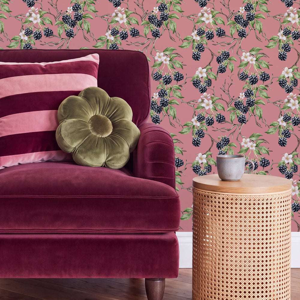 A styled interior scene featuring a deep burgundy velvet armchair with a pink striped pillow and an olive green flower-shaped cushion, set against pink wallpaper with a blackberry and white flower pattern. A small woven side table holds a gray ceramic mug.
