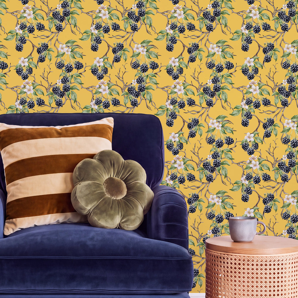 A styled interior scene featuring a mustard yellow wallpaper with a blackberry and white flower pattern, a deep blue velvet armchair with striped and flower-shaped pillows, and a small woven side table with a cup.