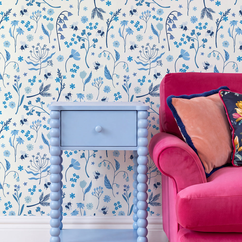 Blue and navy floral wallpaper with delicate botanical illustrations on a white background, styled in a room setting with a light blue bead-frame side table and a bright pink armchair with decorative pillows.