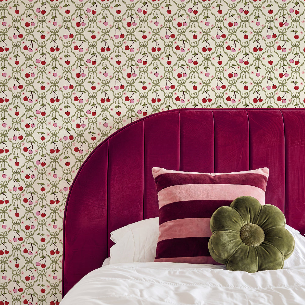 A styled bedroom corner featuring a bed with a deep magenta velvet headboard, white bedding, a pink and burgundy striped pillow, and an olive green flower-shaped pillow. The wall behind is covered in cream wallpaper with a repeating red cherry and green vine pattern.