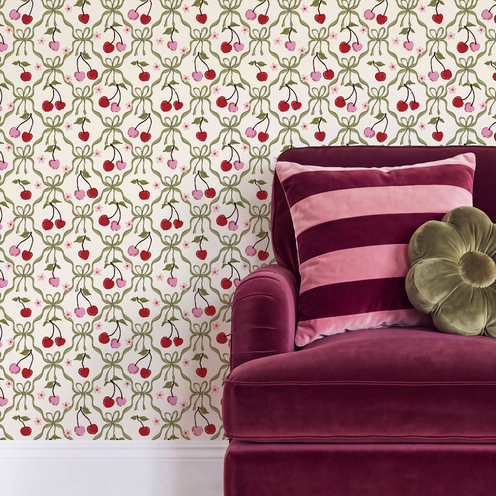 A close-up of a patterned wallpaper featuring red cherries with green stems and pink bows on a cream background, styled behind a deep magenta velvet armchair with pink striped and olive green pillows.