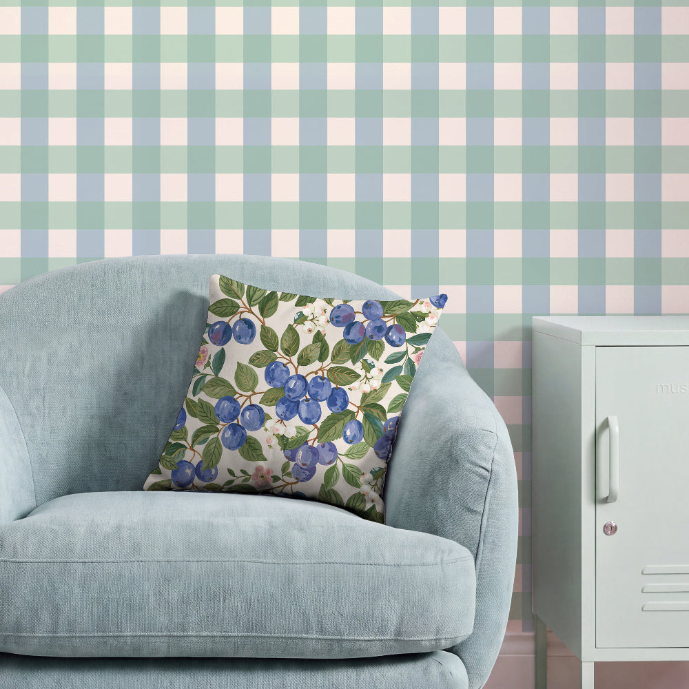 A light blue armchair in a styled room setting, featuring a square throw pillow with a blueberry and green leaf pattern on a cream background. The wall behind has a pastel blue and pink gingham check pattern, and a white cabinet is visible on the right.