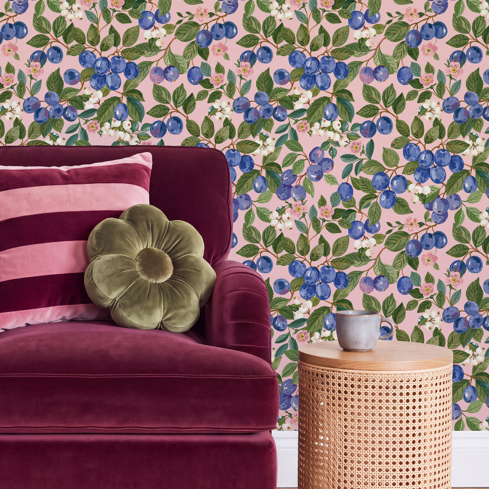 A styled interior scene showing a burgundy velvet armchair with pink striped and olive green flower-shaped pillows, next to a woven rattan side table. The wall behind is covered in pink wallpaper with a blue blueberry and green leaf pattern.